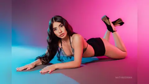 Young woman in black lingerie and heels lying on a colorful pink and blue studio background.