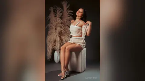 Elegant woman in a white two-piece outfit sitting on a pedestal against a moody, aesthetic pampas grass backdrop.
