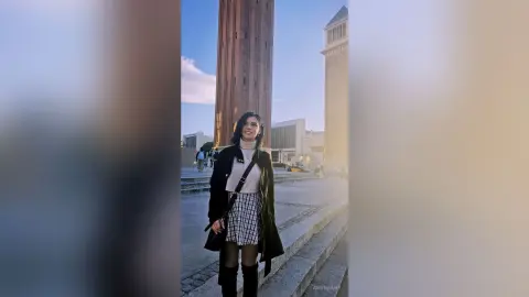 Smiling woman in a black coat and plaid skirt standing in a sunny urban plaza near tall modern buildings.
