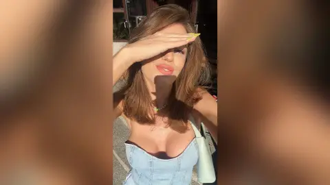 Brunette woman in a light blue denim top posing outdoors in warm sunlight.