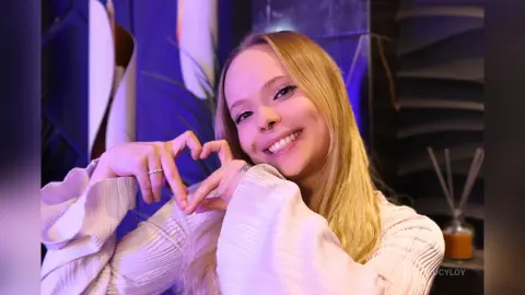 LucyLoy smiles at the camera while making a heart shape with her hands, wearing a white pleated top.