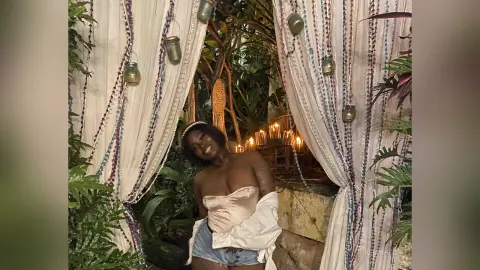 KeianHoz poses naked with a white shirt falling off her shoulders, standing between white curtains in a lush garden setting.