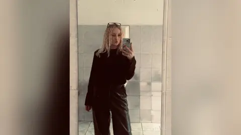 Blonde woman in a black long-sleeve top and leather trousers taking a moody mirror selfie in a tiled bathroom.