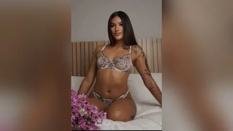 Confident woman in floral lace lingerie sitting on a white bed in a bright, minimalist bedroom.