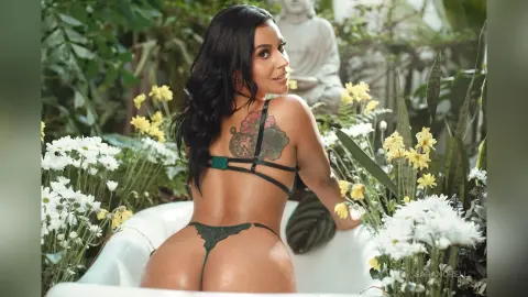 Woman in green bikini sitting in a white bathtub surrounded by white and yellow flowers in a lush garden.