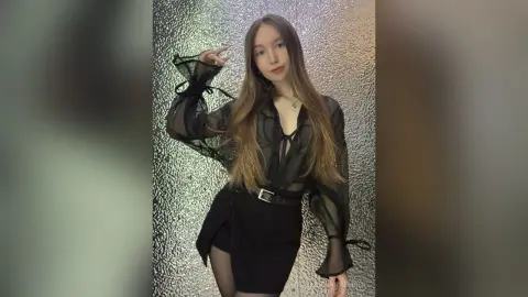 Young woman in a sheer black long-sleeve blouse and mini skirt posing against a shimmering silver backdrop.
