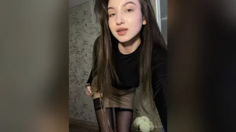 Young woman with long brown hair wearing a black long-sleeve top and tan skirt in a cozy indoor setting.