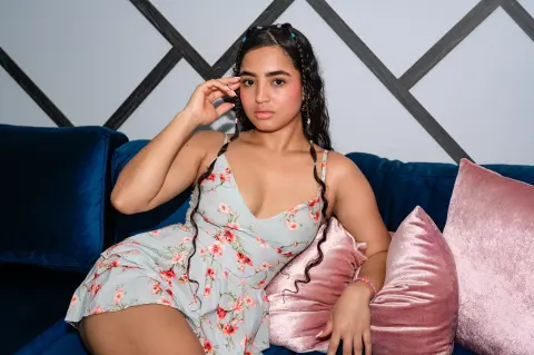 Young woman in a floral summer dress relaxing on a blue velvet sofa with pink pillows in a modern room.