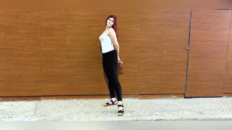 Red-haired woman in a white tank top and black leggings posing against a brown wood paneled wall.