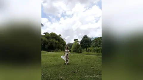 AmelieLautrec stands in a grassy field wearing a long, white, flowing dress with a high slit, surrounded by green trees under a cloudy sky.