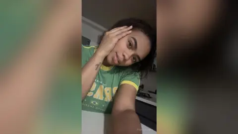PamelaCreed wearing a green t-shirt with yellow trim, touching her hair and sticking her tongue out slightly.