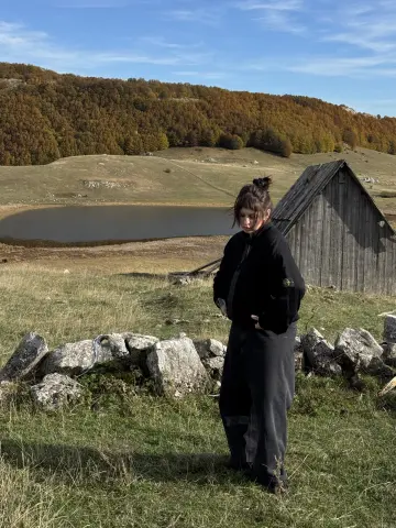AlisaMoss stands in a grassy field near a wooden shack and a lake, wearing a black hoodie and dark pants.