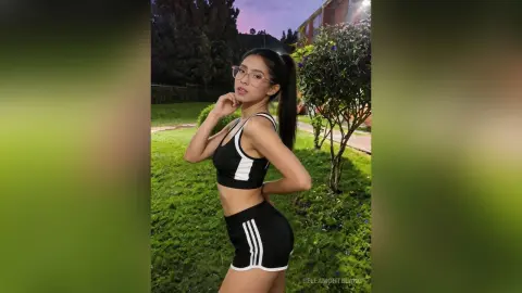 Young woman in black athletic crop top and shorts posing confidently in a lush green garden at twilight.