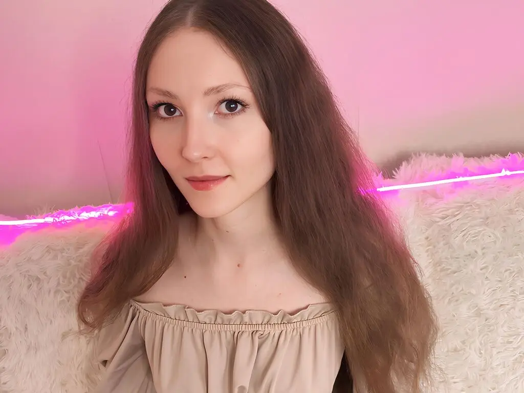 Young woman with long brown hair wearing a beige off-the-shoulder top in a cozy room with pink neon lighting.