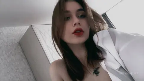 Young woman with long brown hair and red lipstick posing in a cozy white bedroom setting.