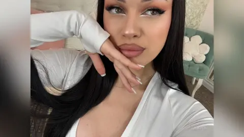 AriaVex poses with her hand against her chin, wearing a white long-sleeved top with long black hair.