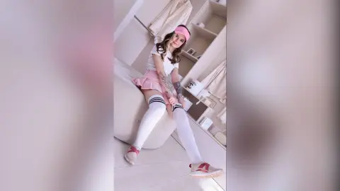 AmeliePearly wears a pink skirt, white top, and white thigh-high socks while sitting on a bed in a light-colored room.