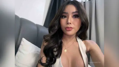 Beautiful woman with long wavy hair wearing a white halter top in a cozy bedroom setting.