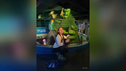 Smiling woman in a white sweater sitting next to a giant Hulk statue at a Marvel themed attraction.