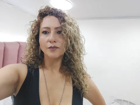 Woman with curly blonde hair wearing a black tank top and gold necklace in a bright room.
