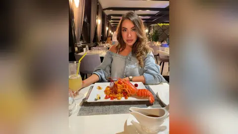 VictoriaWynn sits at a restaurant table wearing a light blue denim shirt, posing with a whole cooked lobster and a yellow drink in front of her.