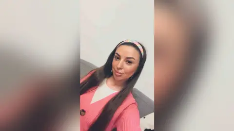 Smiling woman in a pink sweater and colorful headband relaxing on a grey sofa in a bright room.