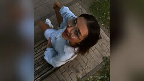 LeonidaAbdon looks up at the camera wearing glasses and a light blue sweatshirt while sitting outside on a wooden bench.