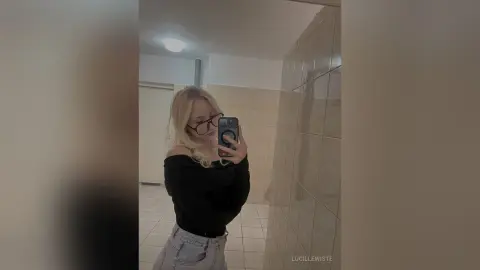 Blonde woman in a black off-the-shoulder top and glasses taking a mirror selfie in a modern bathroom.