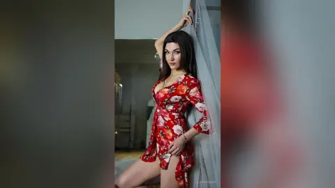 Elegant woman in a red floral wrap dress posing gracefully against a sheer curtain in a bright room.