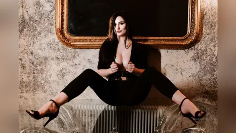 KassieWeins sits on a white radiator wearing a black top and black pants, posing in front of a large gold mirror.