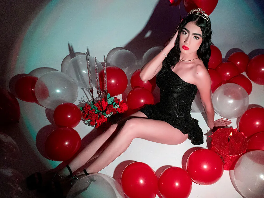 MaluCortes wears a black mini dress and tiara while reclining on a floor covered in red and white balloons.