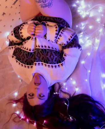 Young woman in a cozy patterned sweater lying on white bedding surrounded by glowing purple fairy lights.