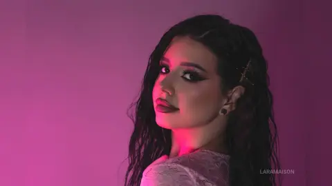 Young woman with dark wavy hair and winged eyeliner posing against a moody pink studio background.