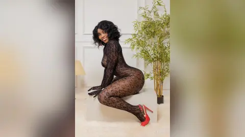Smiling Black woman in a sheer black lace bodysuit and red heels sitting in a bright minimalist room.