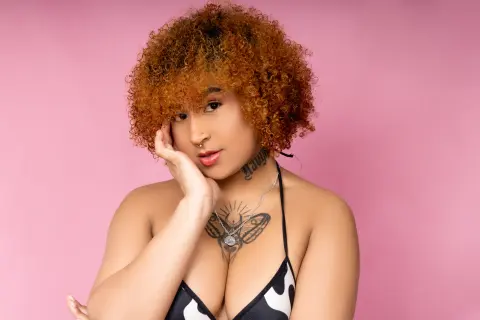 Smiling woman with curly ginger hair and tattoos wearing a black bikini against a pink background.