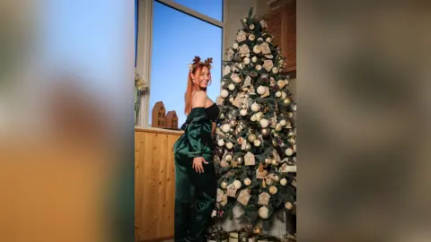 Smiling woman in emerald green satin pajamas standing next to a decorated Christmas tree in a bright room.
