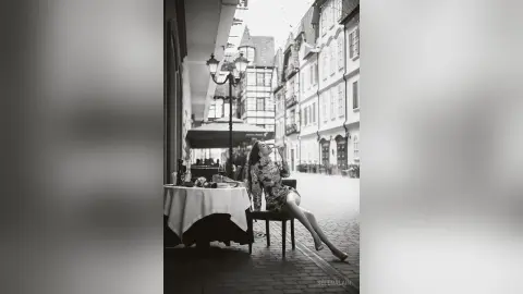 Elegant woman in a patterned dress sipping a drink at an outdoor cafe in a European cobblestone street.