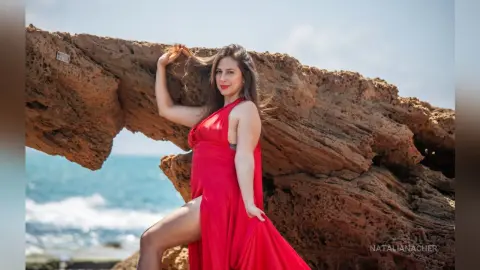 NataliaNacher poses outdoors in a long red dress with a high slit, standing against a large rock formation by the ocean.