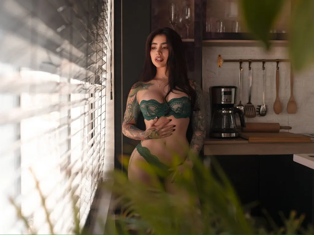 Young woman in green lace lingerie standing by a bright window in a modern kitchen setting.