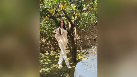 Elegant woman in a white lace bodysuit and trousers standing under a sunny lemon tree in a rustic garden.