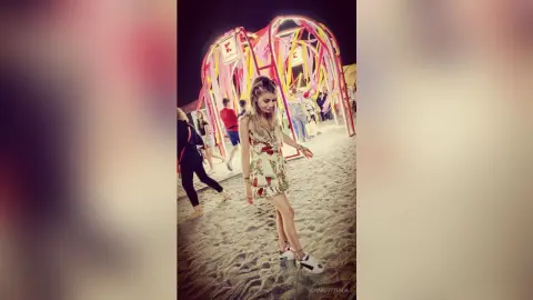 Young woman in a floral sundress standing on a sandy beach at night near a colorful pink decorative installation.