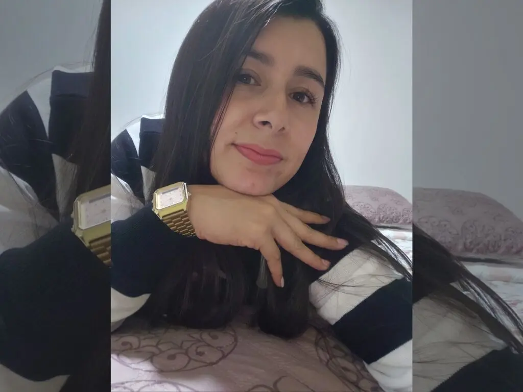 Young woman with long dark hair wearing a white and black sweater relaxing on a bed in a cozy indoor setting.
