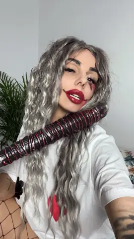 Woman in Harley Quinn cosplay with wavy grey hair and red makeup holding a prop bat in a bright room.