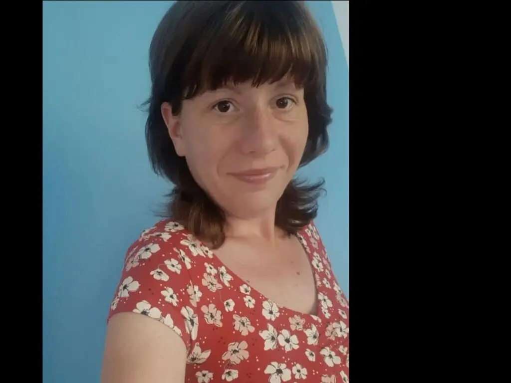 AliceKatty smiles at the camera, wearing a red floral print t-shirt against a plain blue wall.