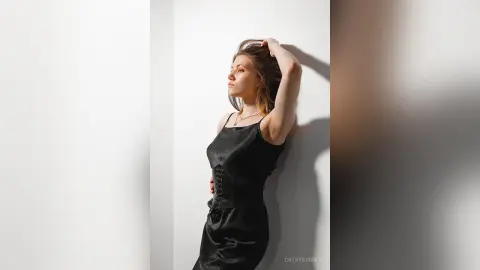 Elegant woman in a black satin slip dress posing against a white wall in soft, moody lighting.