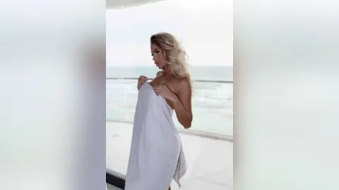 Blonde woman wrapped in a white towel standing on a balcony overlooking the ocean at sunset.