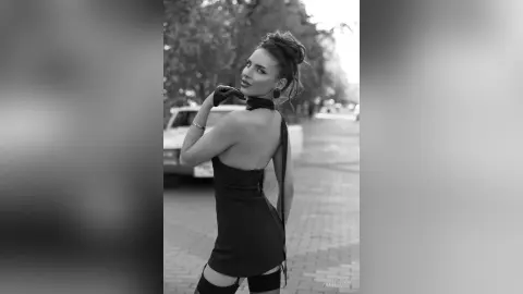 AnjeNoir poses in a short black dress and gloves, looking over her shoulder on a city street.