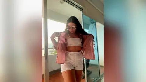 Stylish young woman in a pink shirt and white crop top outfit posing on a sunny balcony.