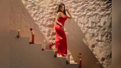 Elegant woman in a long red evening gown posing on stairs by glowing candles in a warm, moody setting.