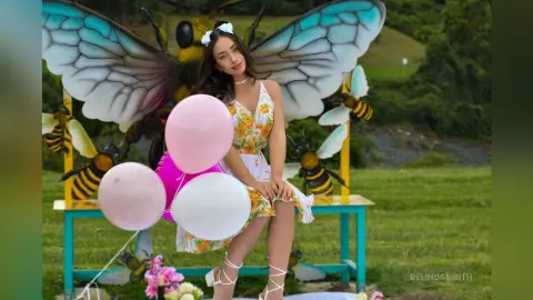 Woman in a floral sundress sitting by giant bee decorations with pink and white balloons in a garden setting.